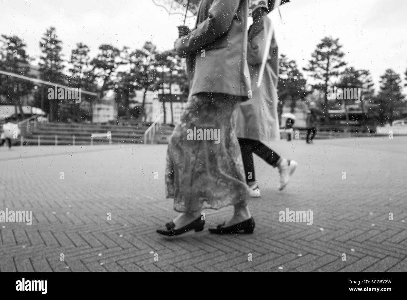 Zwei Personen gehen durch einen regnerischen Stadtplatz, in Schwarz-weiß gefangen, was Ruhe und Bewegung vermittelt. Regentropfen auf der Linse verstärken die Ruhe Stockfoto