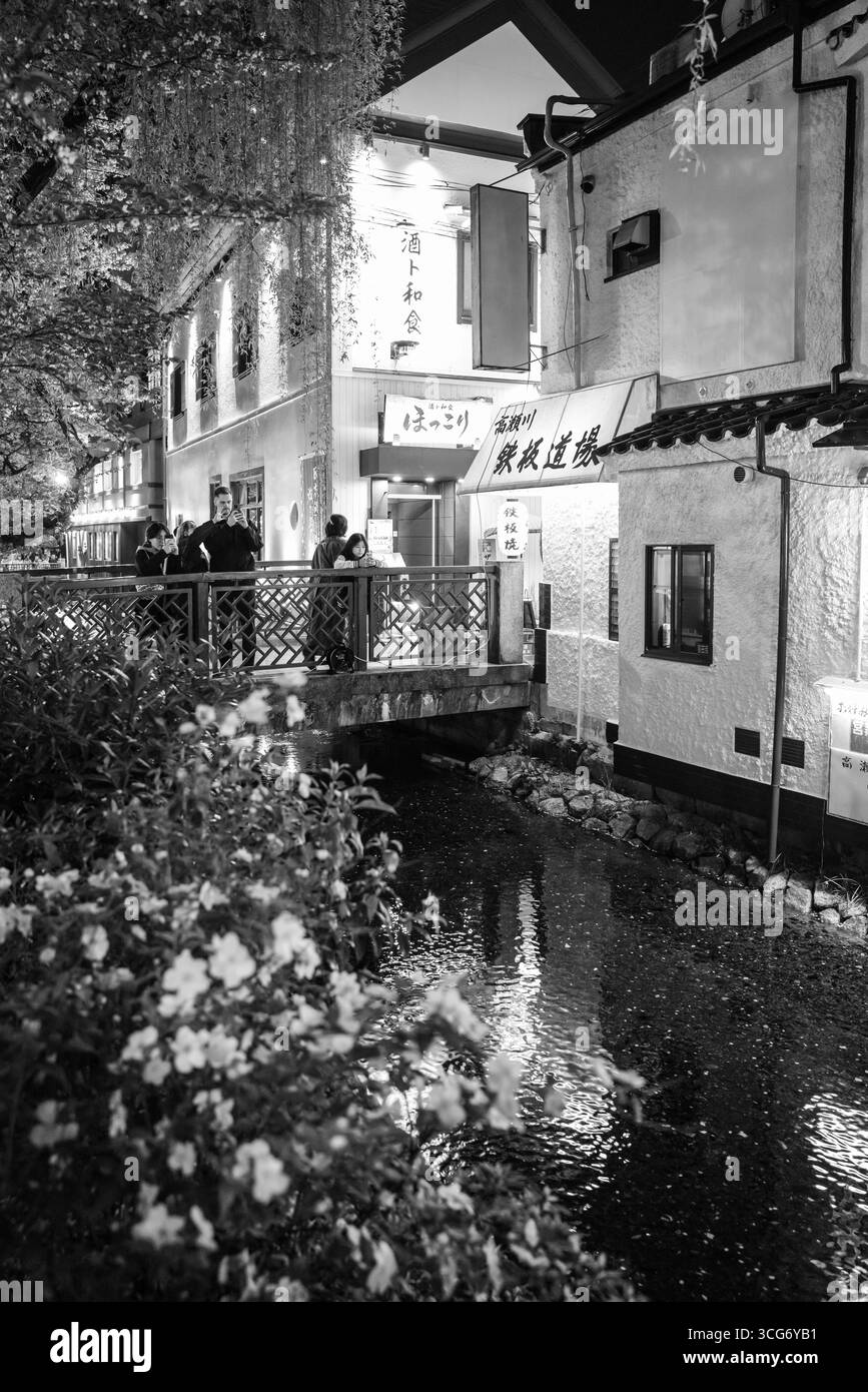 Schwarzweiß-Foto einer traditionellen japanischen Straße bei Nacht mit dem Takase River und wunderschönen blühenden Sakura-Bäumen. Die Lage ist ruhig Stockfoto