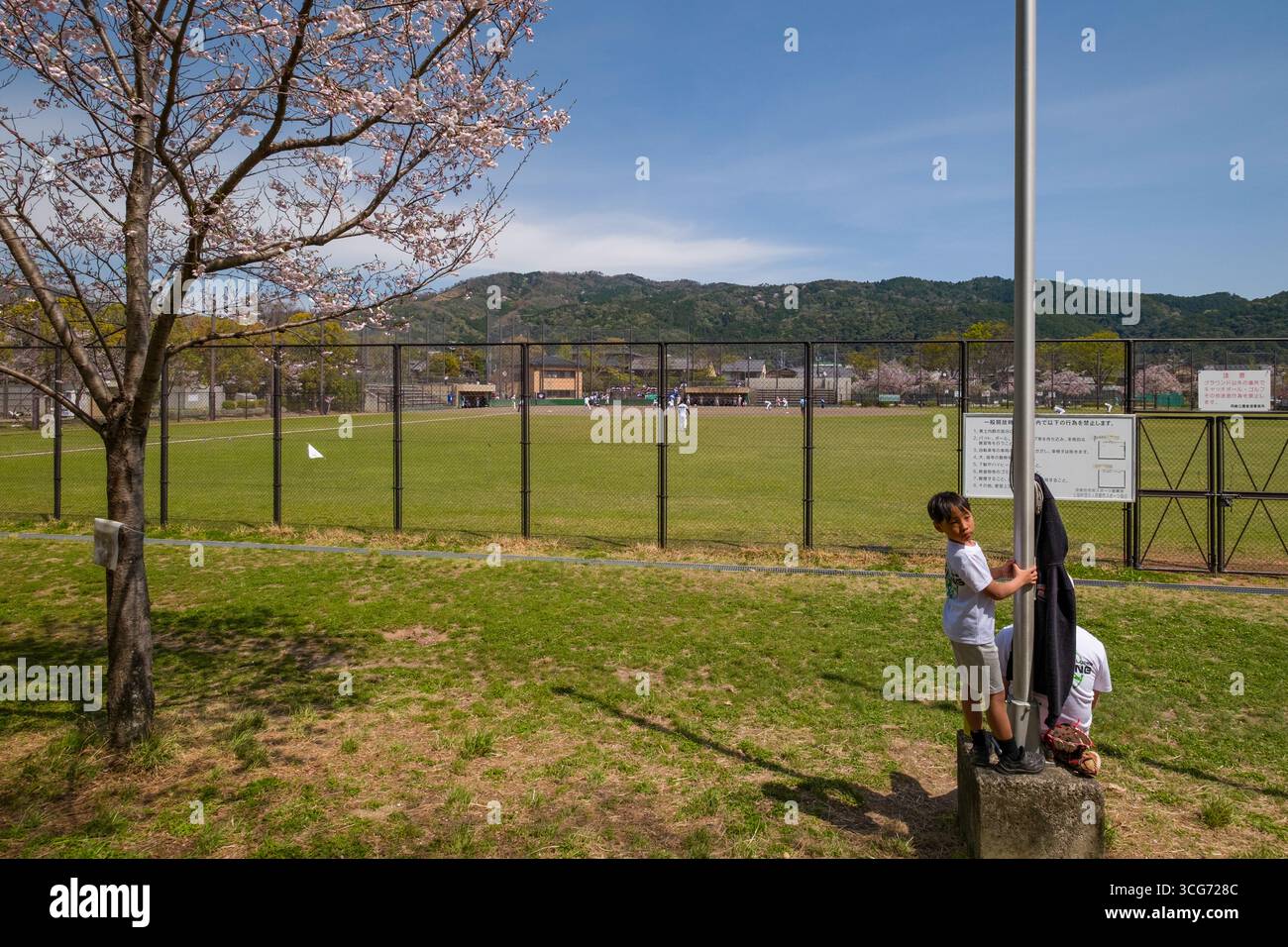 Junge, der auf Betonsockel einer Lampenstange mit Baseballfeld im Hintergrund im Okazaki Park, Okazaki Saishojicho, Sakyo Ward, Kyoto, Japan steht Stockfoto