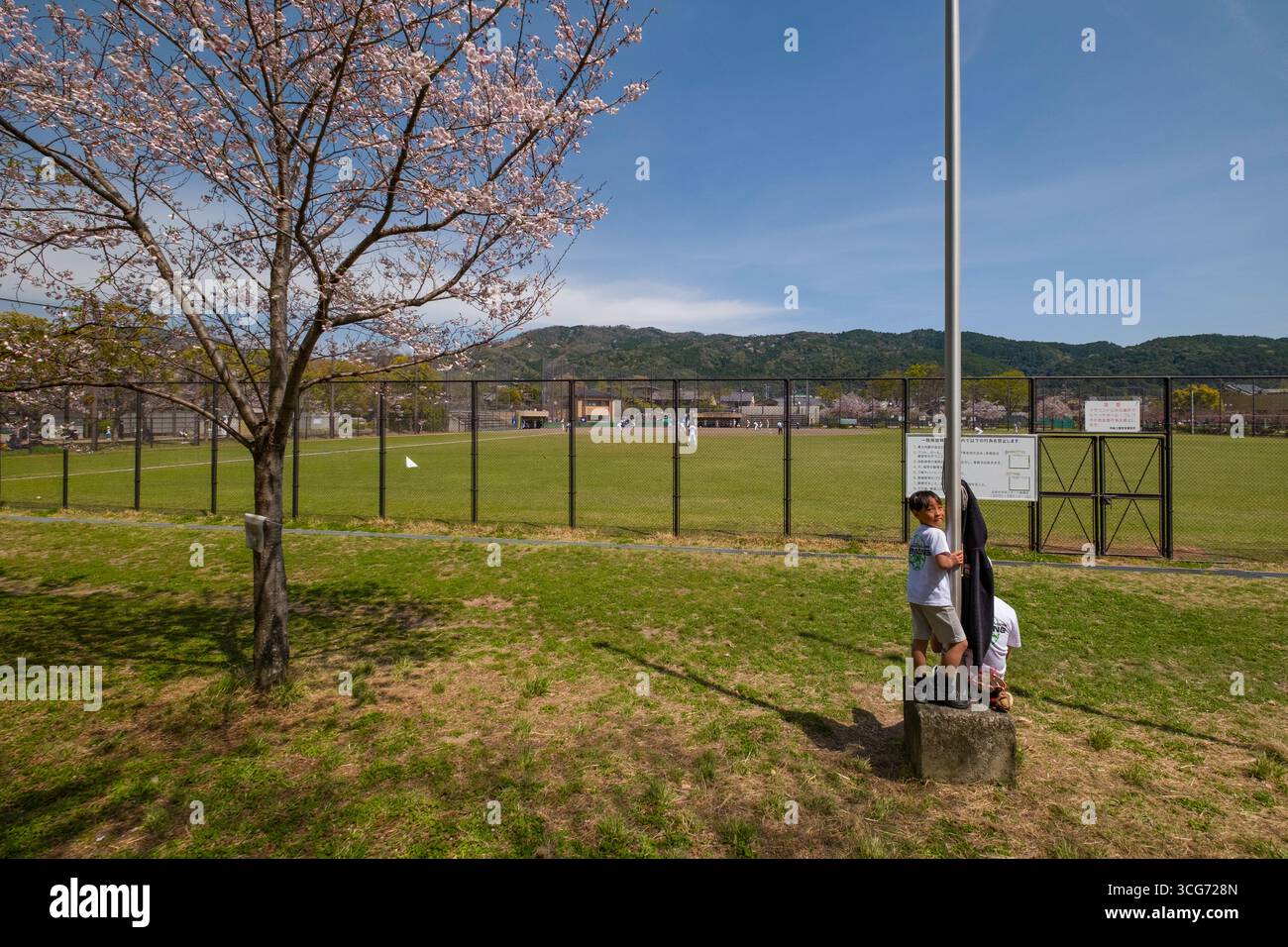 Junge, der auf Betonsockel einer Lampenstange mit Baseballfeld im Hintergrund im Okazaki Park, Okazaki Saishojicho, Sakyo Ward, Kyoto, Japan steht Stockfoto