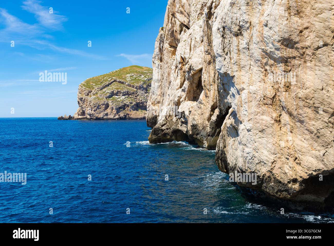 Helle Kalksteinklippen von Capo Caccia, Sardinien, die sich über tiefblauem Meer mit klarem Sommerhimmel und weit entfernten felsigen Vorgebirgen erheben. Stockfoto