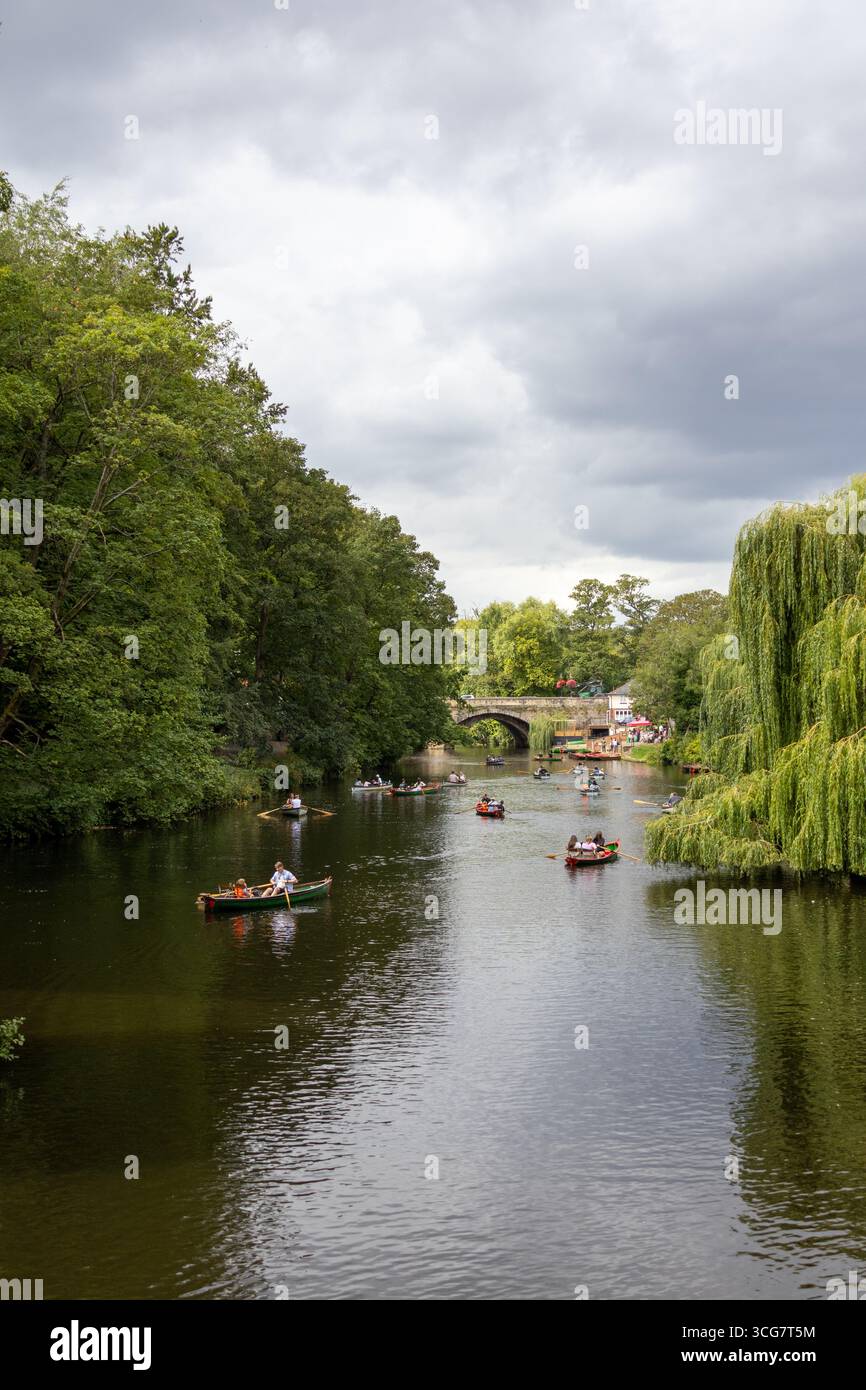 Malerische Aussicht auf Menschen, die auf dem Fluss Nidd in Knaresborough, North Yorkshire, rudern, mit einer Steinbrücke und dichten grünen Bäumen unter bewölktem Himmel Stockfoto