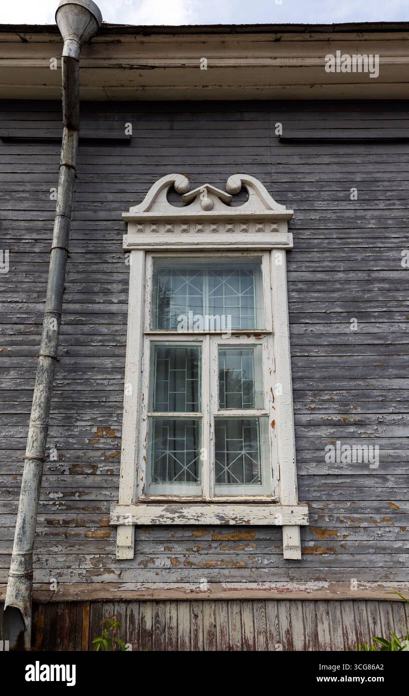 Das alte Holzhaus präsentiert ein einzelnes, weiß gerahmtes Fenster mit dekorativen Zierleisten, veralteter Textur und rustikalem Charme des russischen Dorfes Stockfoto