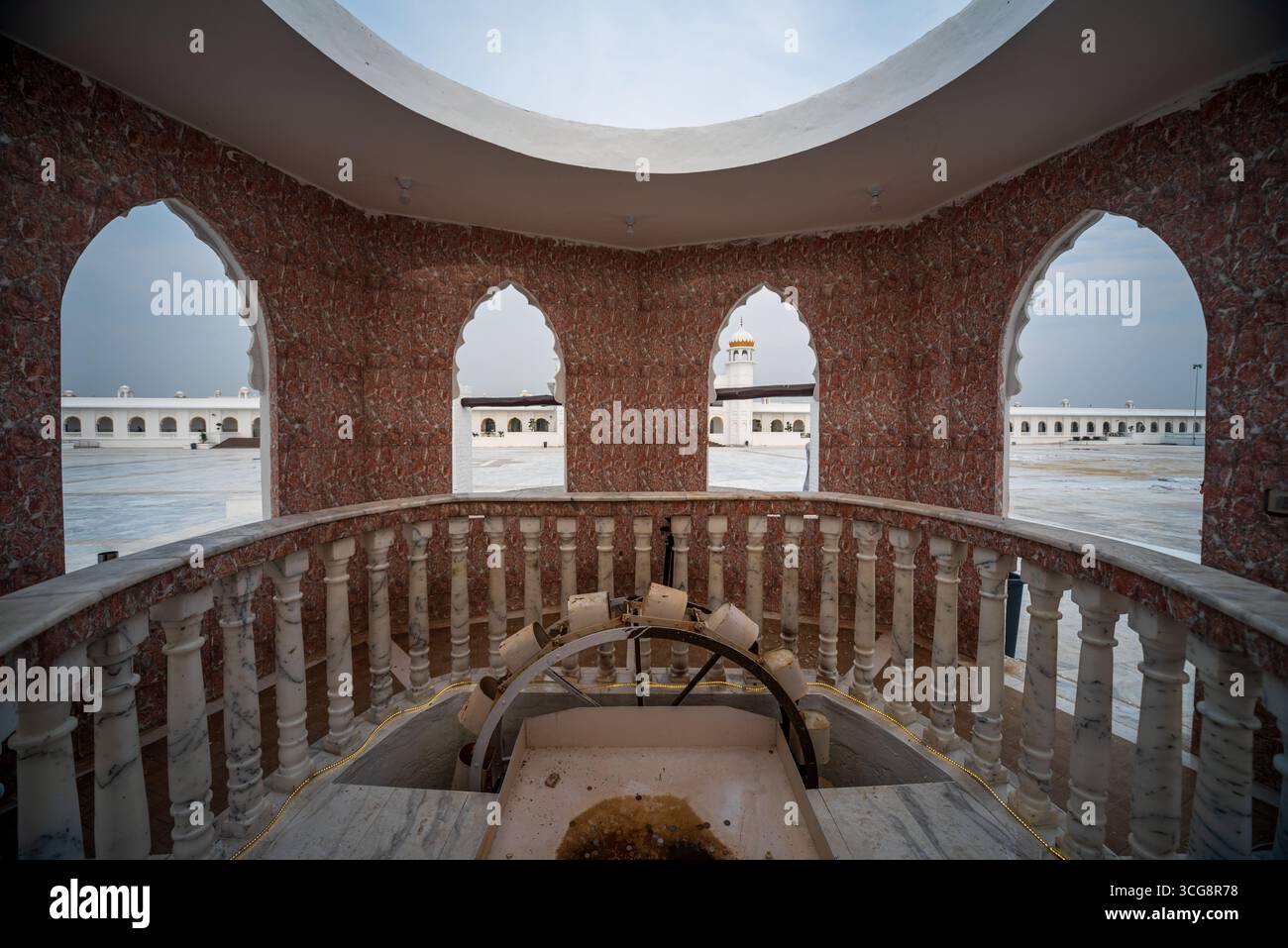Blick auf die kunstvoll verzierte Marmorstruktur mit Bogenfenstern, die die entfernten weißen Gebäude unter einem weichen Himmel umrahmen, eine Mischung aus Tradition und Gelassenheit, Kartarpur, Punjab, Pakistan. Stockfoto