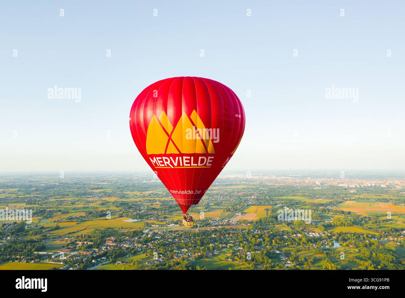 Sint-Martens-Latem, Belgien - 25. August 2025: Aus der Vogelperspektive eines leuchtenden roten Heißluftballons, der über der üppigen grünen Landschaft schwingt, ein Leuchtfeuer vor dem weichen, blauen Himmel. Stockfoto