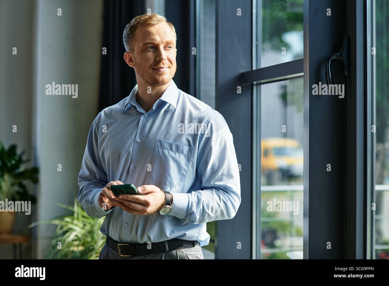 Ein geschickter Geschäftsmann steht in einem Büro, hält ein Smartphone in der Hand und blickt nachdenklich nach draußen. Stockfoto