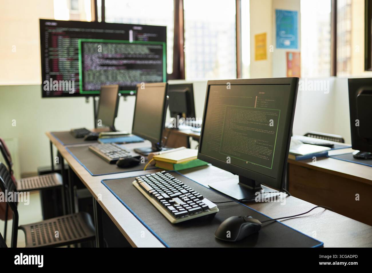 Leeres Computerlabor mit mehreren Desktop-Monitoren, die Programmiercode auf Bildschirmen anzeigen, Tastaturen und Mäuse auf Schreibtischen angeordnet, großes Fenster mit natürlichem Licht, keine Personen anwesend Stockfoto