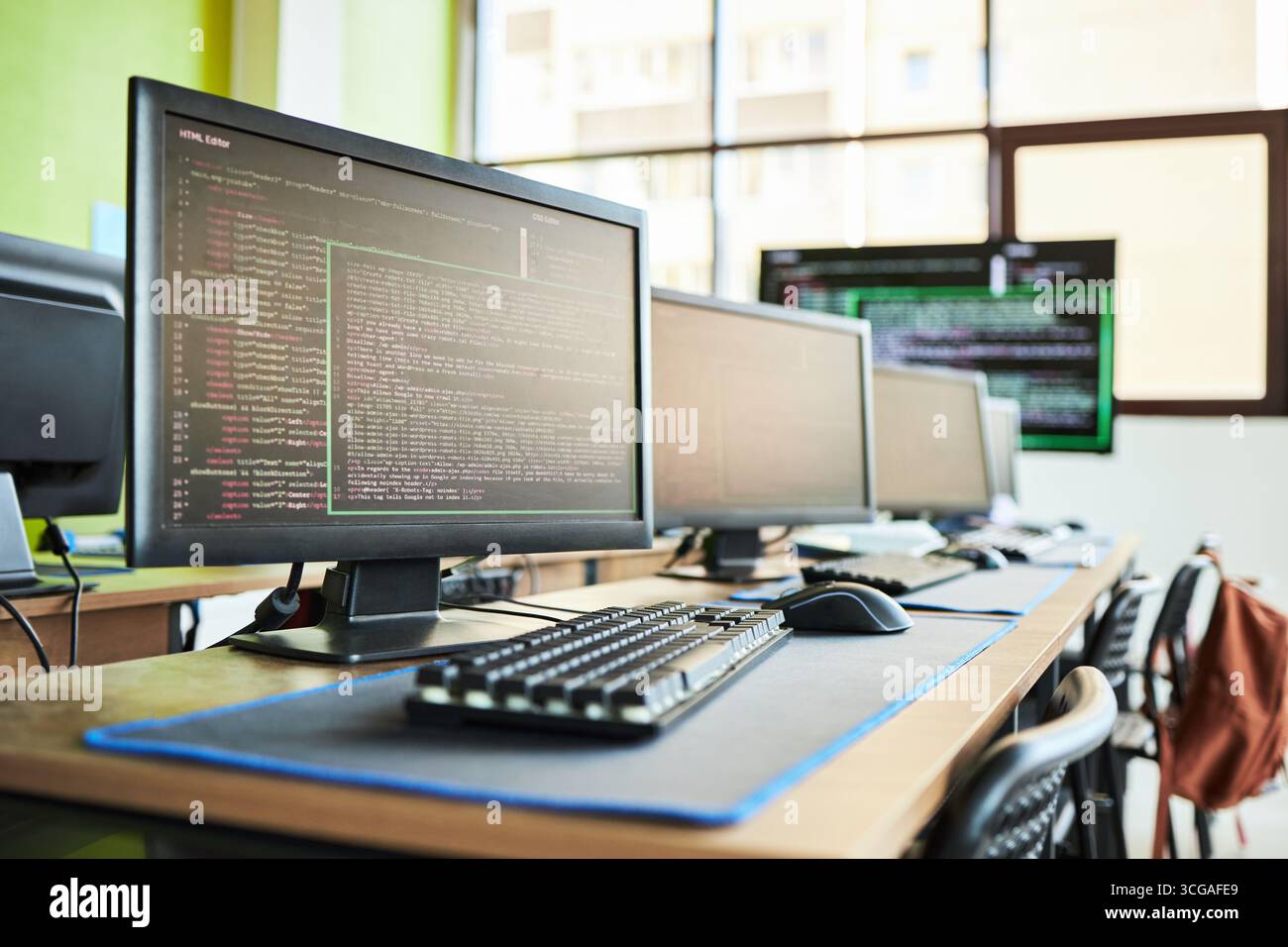 Reihe von Desktop-Computern, die Programmiercode auf Monitoren anzeigen in modernen Computerlabors, leere Stühle an Schreibtischen, Tastaturen und Mäuse, die ordentlich angeordnet sind, heller Fensterhintergrund Stockfoto