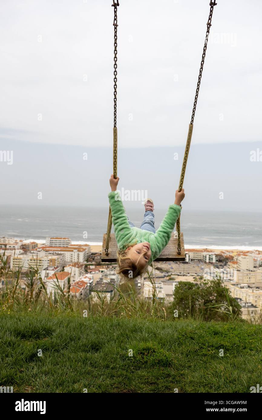 Fröhliches Kind schwingt in der Nähe der Küstenstadt, Sonnenlicht schimmert über die Wellen des Ozeans und die urbane Landschaft Stockfoto