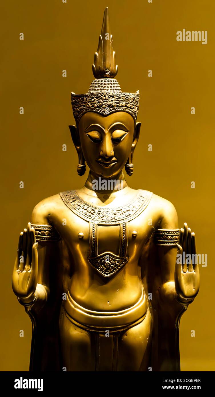 Eine goldene Statue einer Buddha-Figur mit einem ruhigen Ausdruck, die Hände in einer Geste des Friedens erhoben, vor einem warmen gelben Hintergrund Stockfoto
