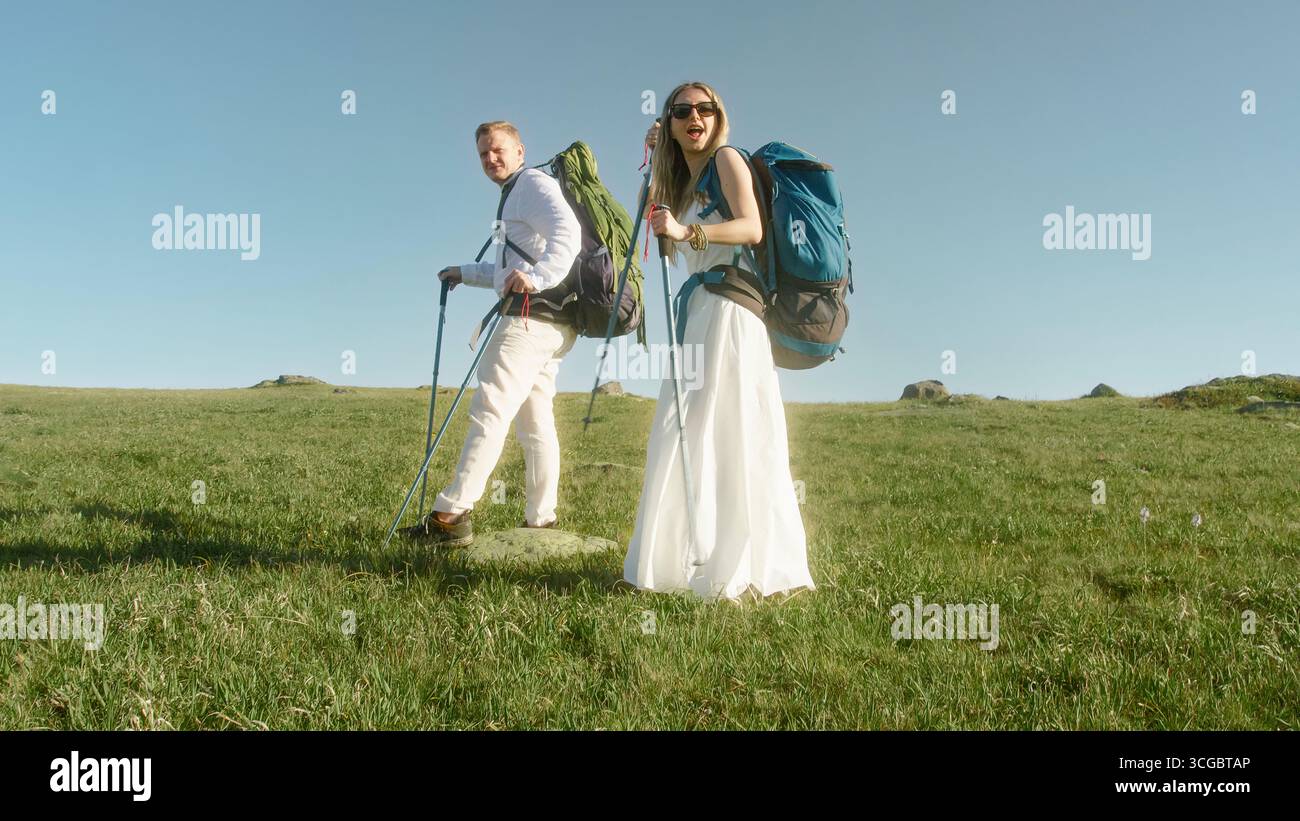 Frisch vermähltes Paar in Hochzeitskleidung, Wandern durch offenes Grasfeld mit Trekkingstöcken und Rucksäcken unter klarem Himmel. Stockfoto