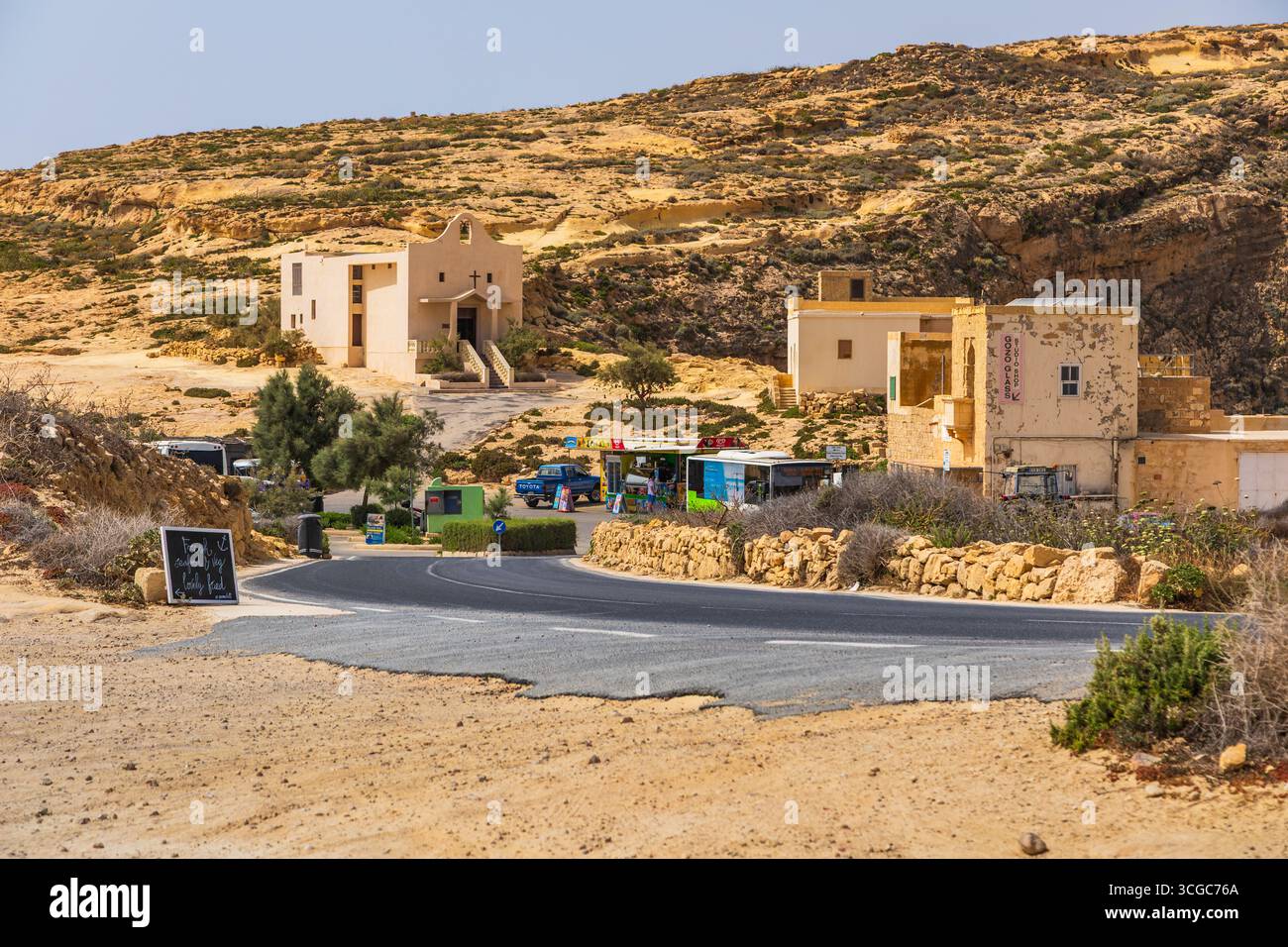 Gozo, Malta – 23. Juni 2021: Gekrümmte Straße, die zu einer kleinen Kapelle und Steinhäusern mit lokalen Imbissständen und Geschäften führt, umgeben von felsigen Hügeln und Ar Stockfoto