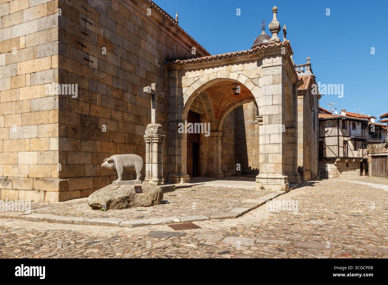 La Alberca, Spanien . 5-31-2024: Steinstatue eines Schweins zu Ehren des Marrano von San Antón und Steinkreuz neben der Kirche unserer Lieben Frau von Assu Stockfoto