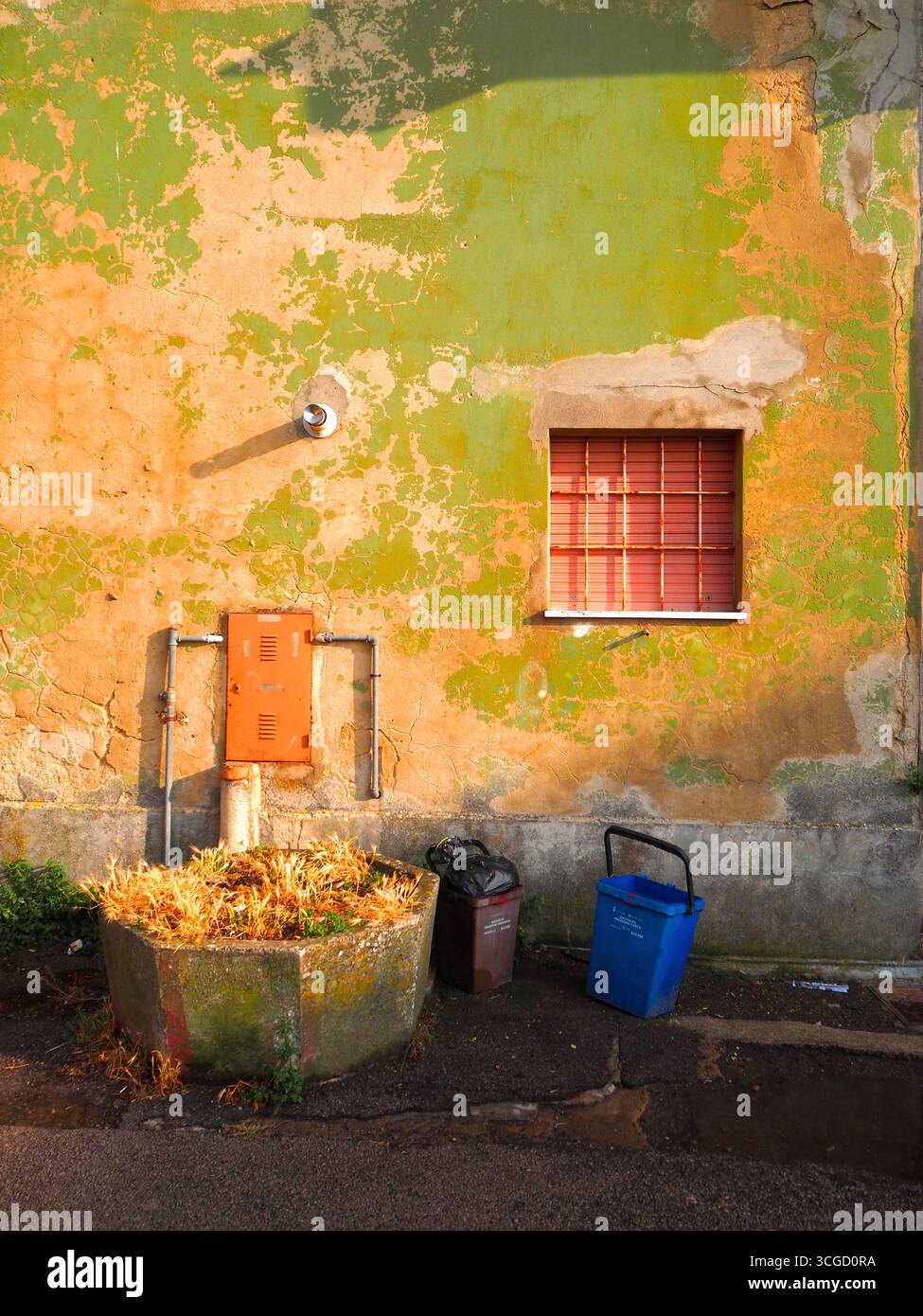 Enna-Wand mit abblätternder grüner und oranger Farbe, beleuchtet durch warmes Sonnenuntergangslicht, rotes Fensterläden, Mülltonnen und kleiner Übertopf in rustikalem Sizilianisch Stockfoto