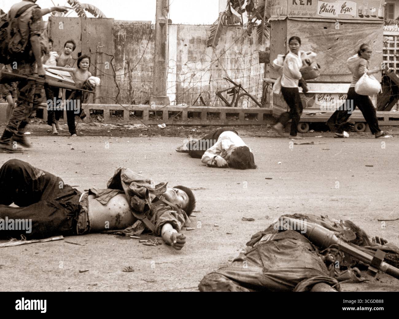 Mit Angst und Besorgnis in ihren Gesichtern und auf Drängen der südvietnamesischen Truppen schleicht sich Frauen und Kinder, die mit geborgenen Besitztümern beladen sind, an den Leichen von drei Vietcong vorbei, die in den Kämpfen getötet wurden. Mai 1968. Stockfoto