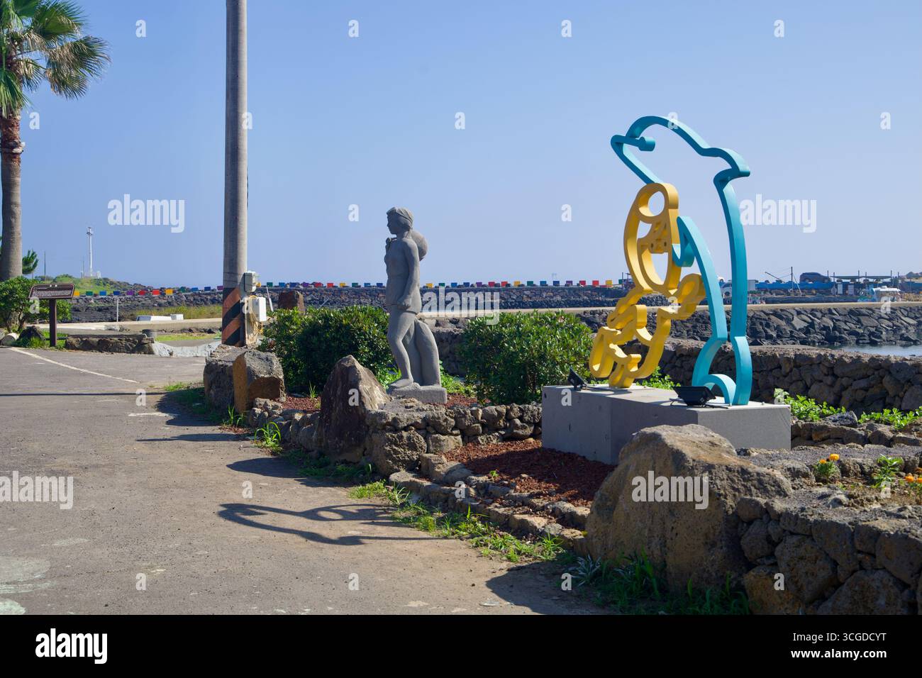 Öffentliche Kunstwerke, darunter eine stilisierte Delfinskulptur und eine Steintaucherfigur, säumen den Hafenweg am Hafen Dongil‑ri mit den farbenfrohen Wellenbrechern Stockfoto