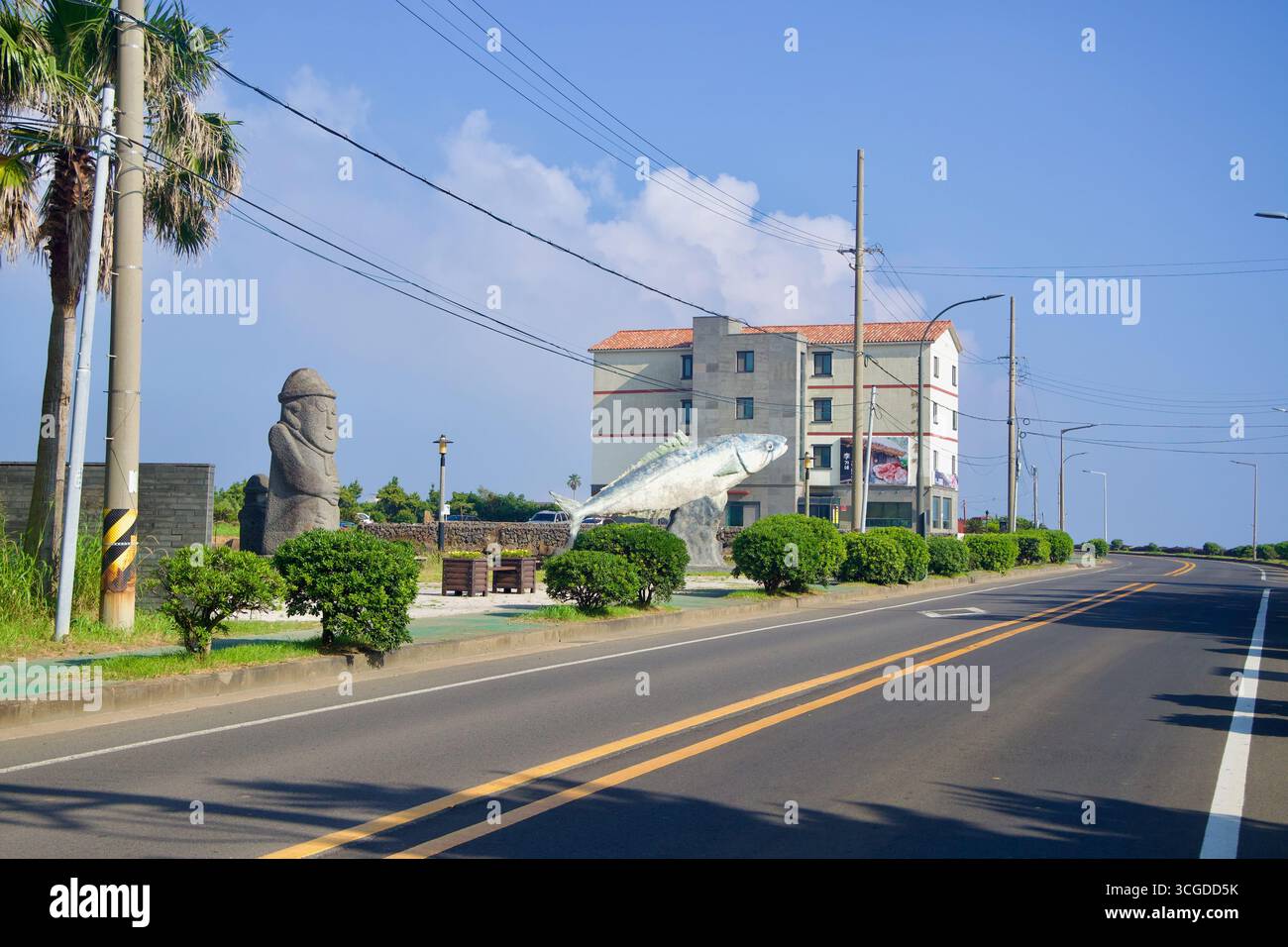 Ein plaza am Straßenrand in der Nähe des Hafens Moseulpo ist mit einer Steinstatue von Dol Hareubang und einer überdimensionalen Fischskulptur neben Hecken und Palmen entlang eines ruhigen C versehen Stockfoto