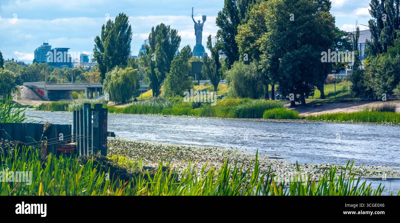 Üppige grüne Vegetation am Fluss Dnipro mit dem Mutter-Ukraine-Denkmal und der Skyline der Stadt Kiew im Hintergrund an einem sonnigen Sommertag Stockfoto