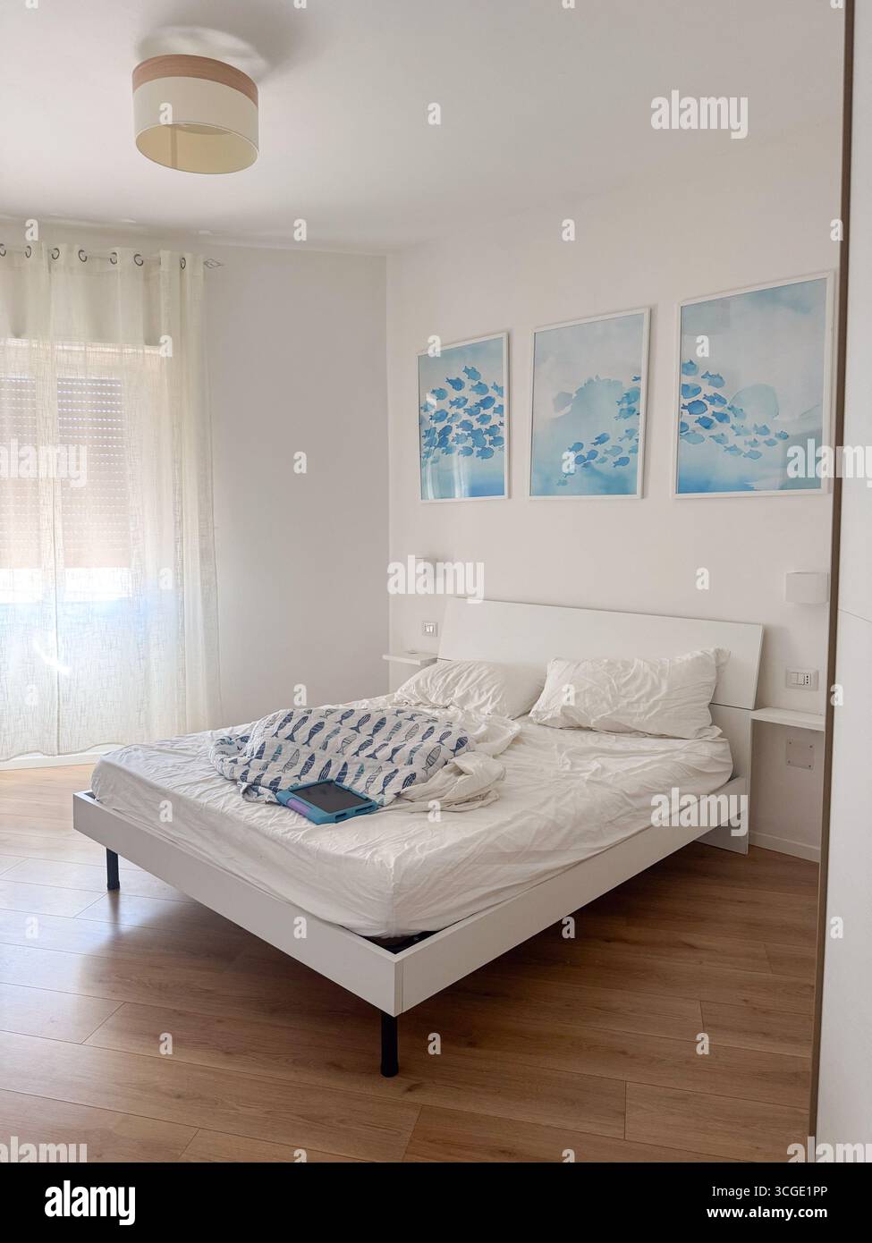 Minimalistisches Schlafzimmer mit ungebautem Bett, einfacher Einrichtung und Holzfußboden. Drei gerahmte Kunstwerke hängen über dem Bett und verleihen der Reinigung Farbe und Stil Stockfoto