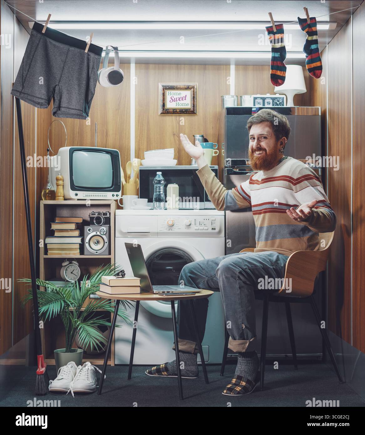 Porträt eines glücklichen, alleinstehenden jungen Mannes, der in einer Mikrowohnung lebt: Er trinkt Kaffee und lächelt in die Kamera Stockfoto