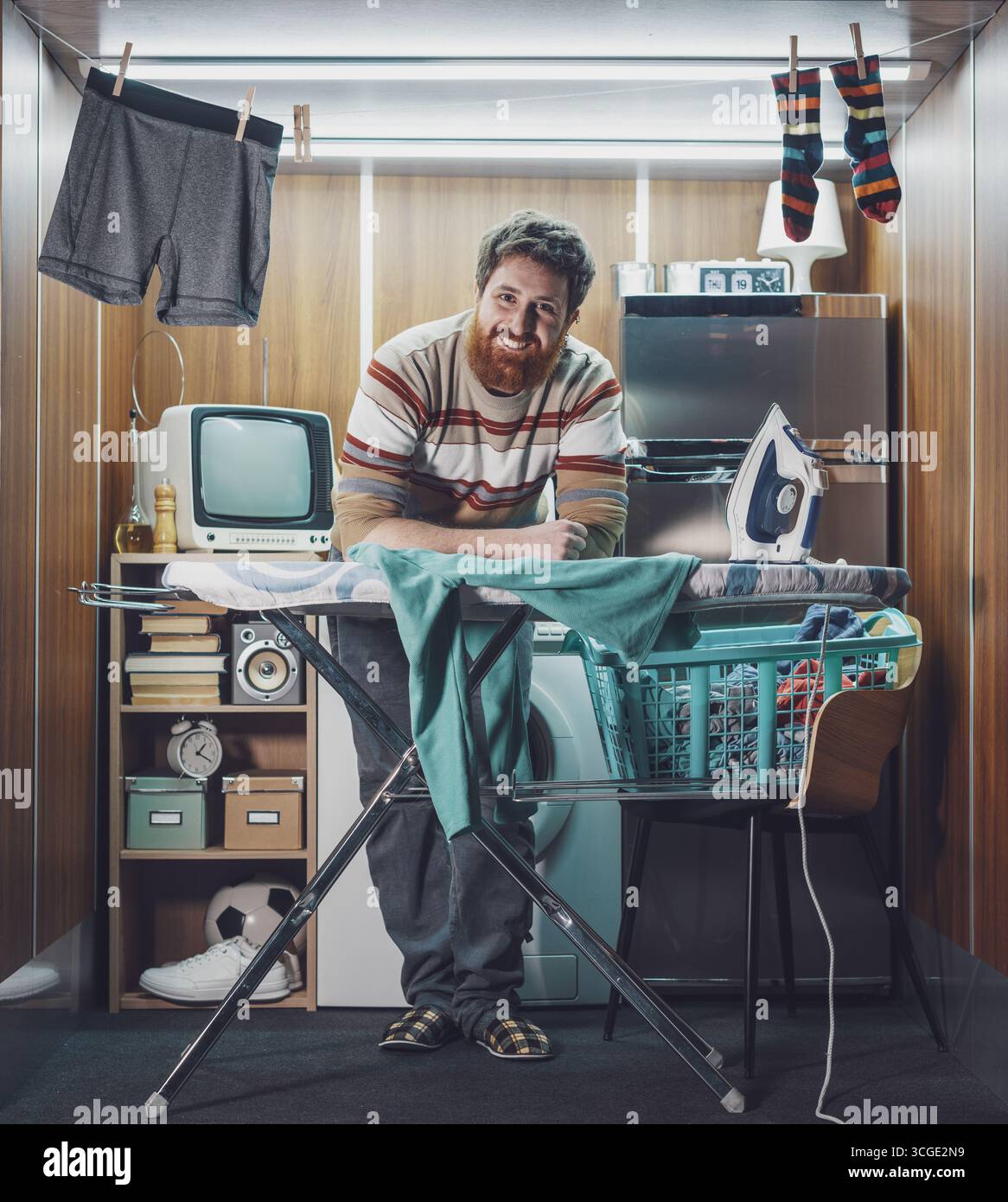 Unabhängiger Mann, der allein in einer Mikrowohnung lebt, bügelt Kleidung und lächelt in die Kamera Stockfoto
