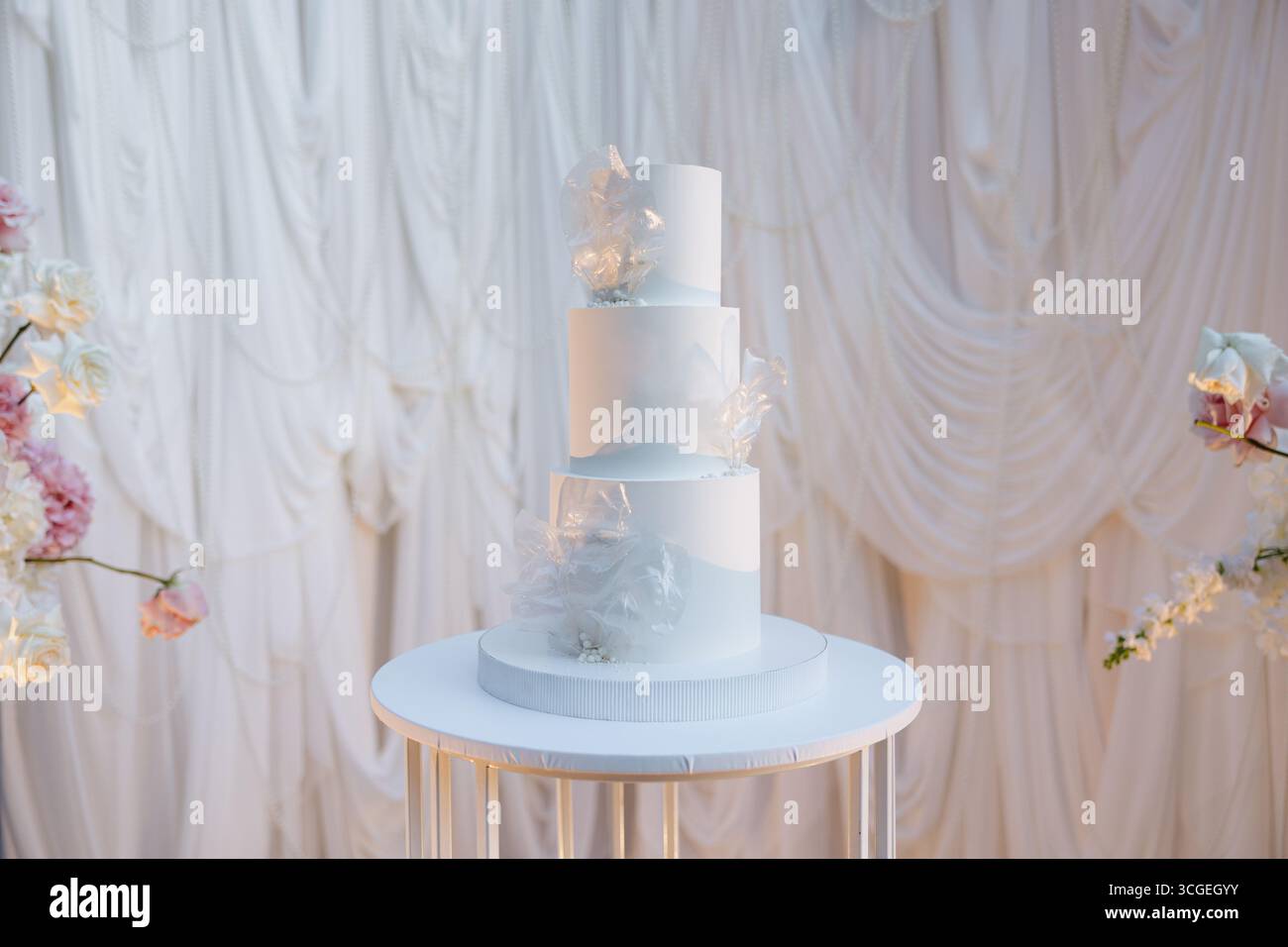 Ein atemberaubender dreistufiger Kuchen steht elegant auf einem Tisch, umgeben von weichem, drapiertem Stoff und Blumenarrangements, die einen romantischen und festlichen Abend einfangen Stockfoto