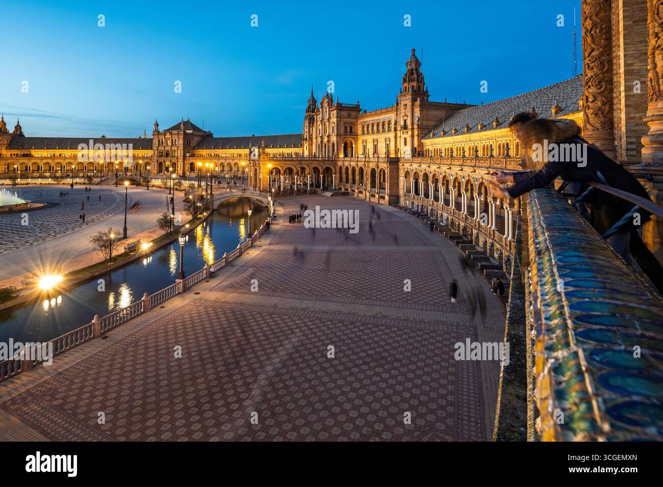 Besucher genießen einen gemütlichen Spaziergang entlang des Kanals der Plaza de España, während warme Lichter die Bögen und Türme in der Abenddämmerung erleuchten. Stockfoto
