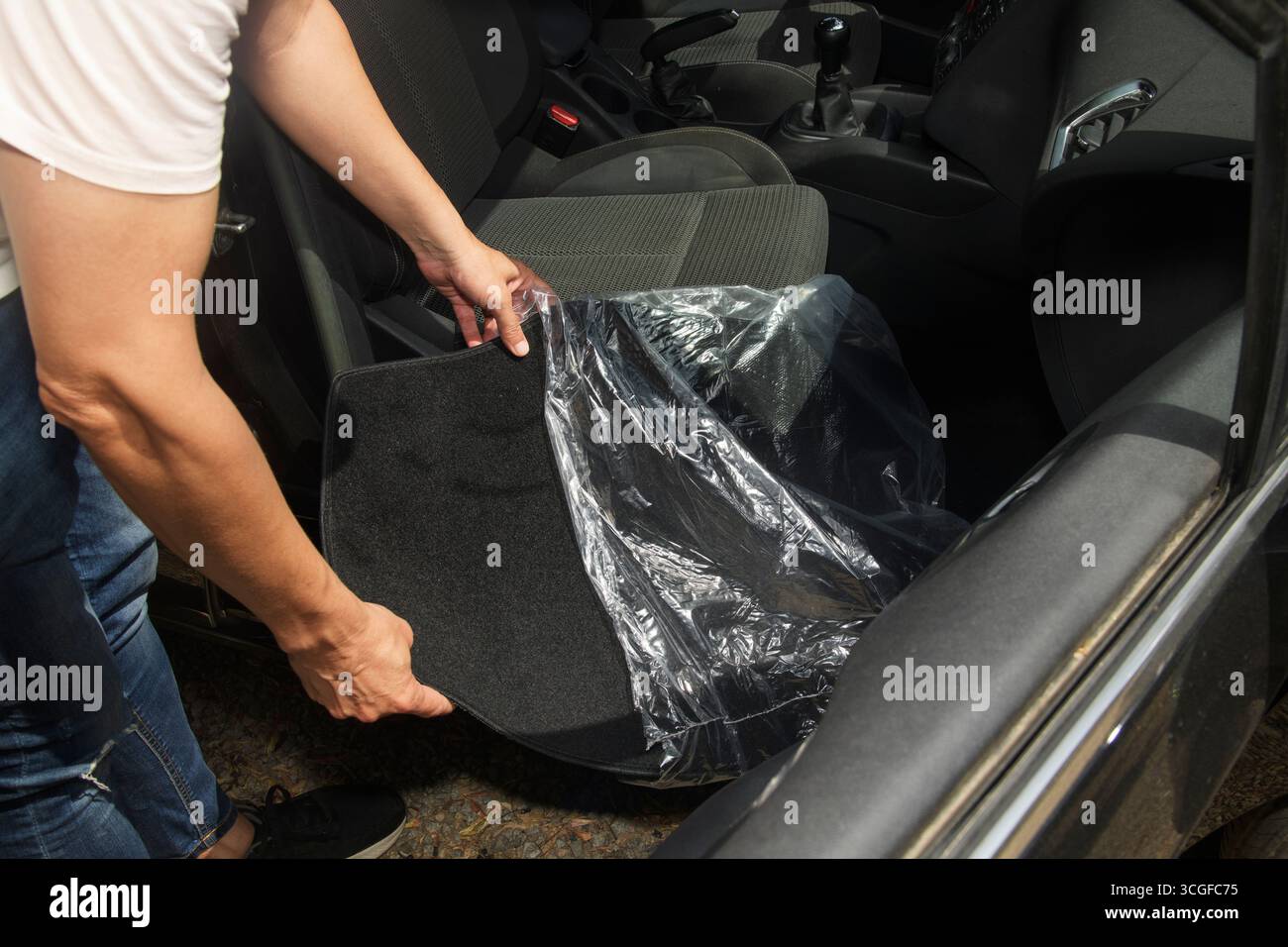 Ein Mann entfernt die Schutzabdeckung aus Kunststoff von einer neuen schwarzen Bodenmatte, bevor sie auf der Beifahrerseite eines Fahrzeuginnenraums platziert wird, um die Montage vorzubereiten Stockfoto