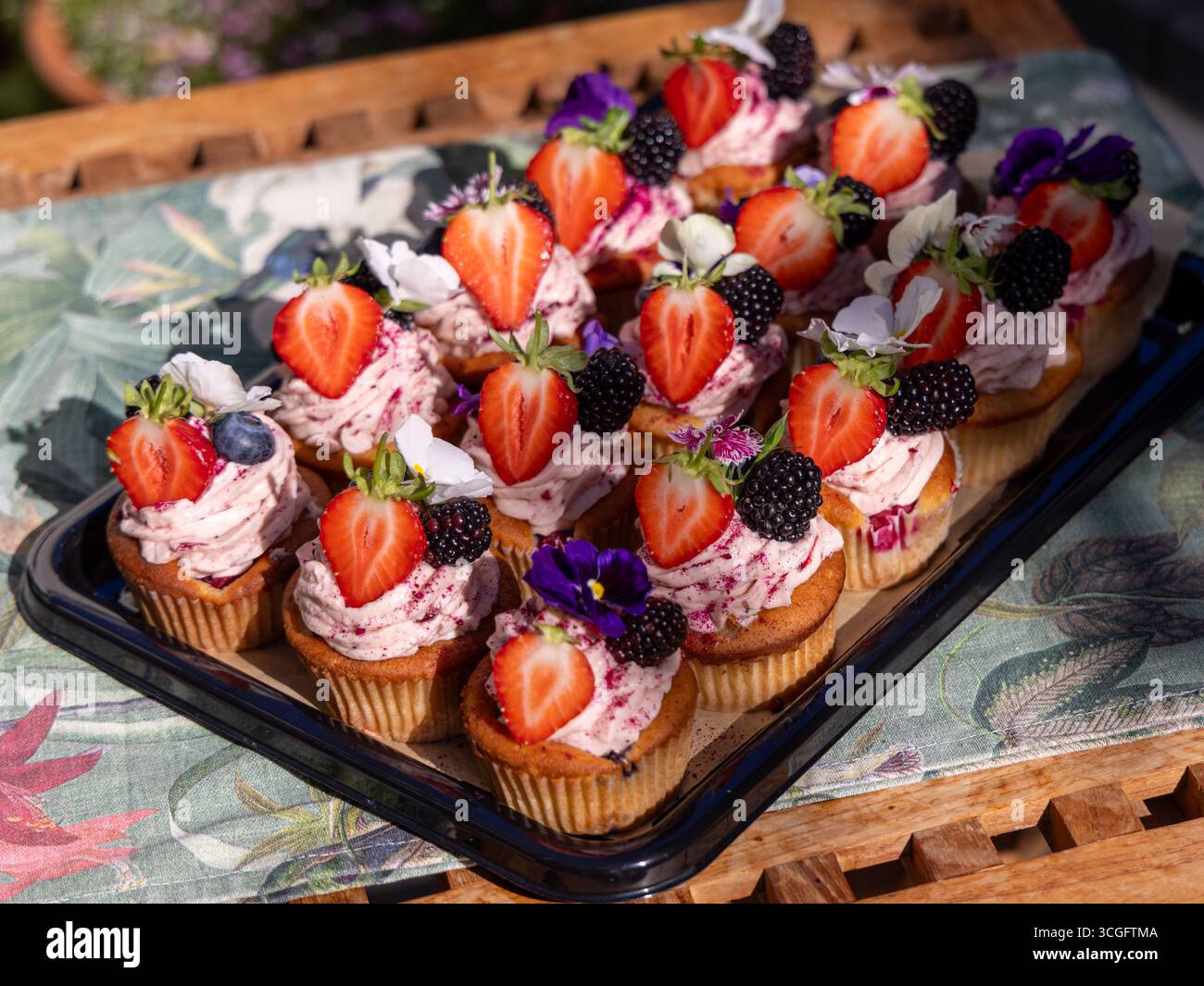 Peggy Porschen Sommerbeeren-Cupcakes auf Tablett mit rosa Zuckerguss, frischem Obst und essbaren Blumen. Ein WhingEats-Gartenmoment. Stockfoto