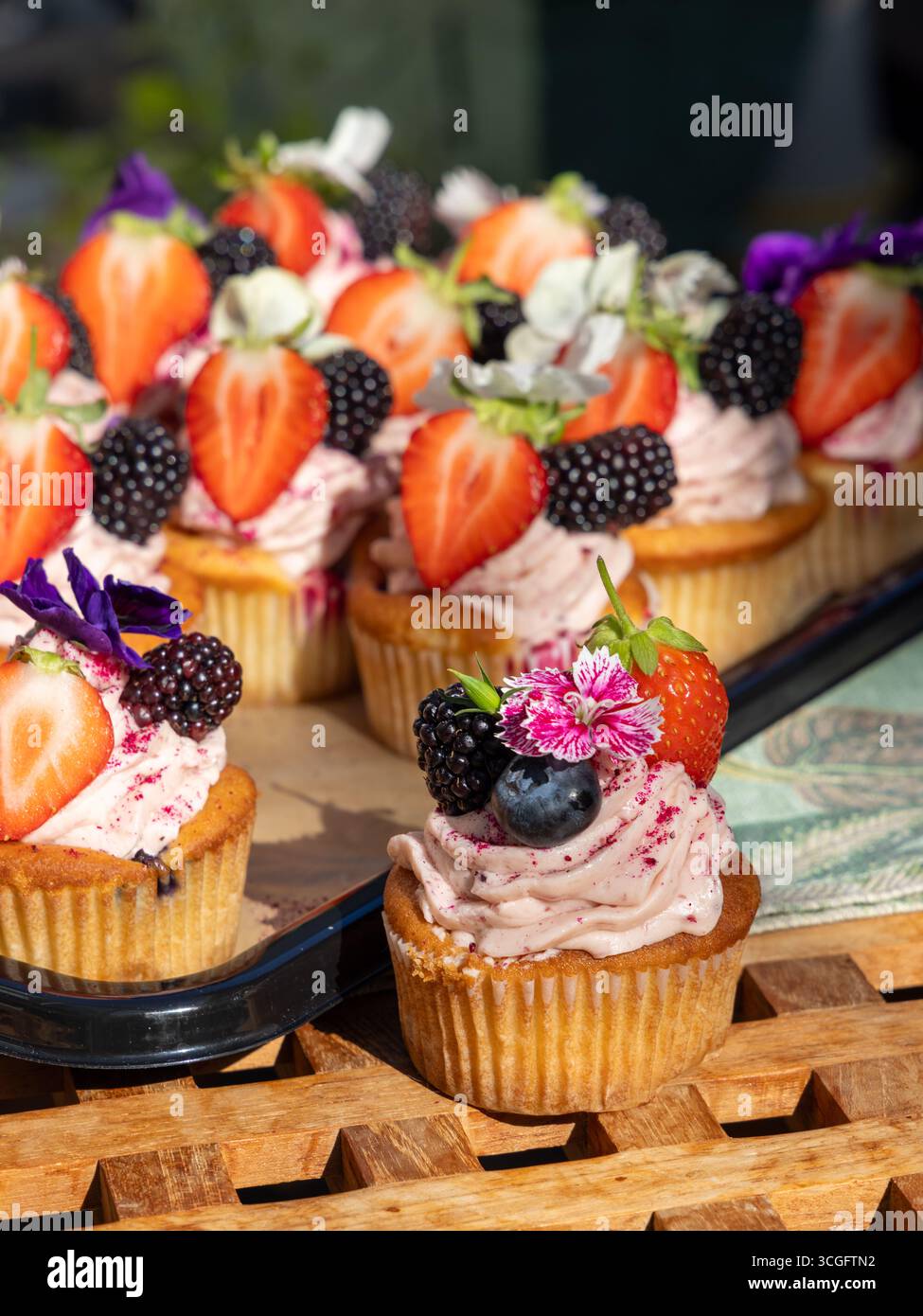 Tablett mit Cupcakes mit rosa Zuckerguss, belegt mit Erdbeeren, Brombeeren, Heidelbeeren und weißen essbaren Blüten auf Holzoberfläche. 🔍 47 Optimiert Stockfoto