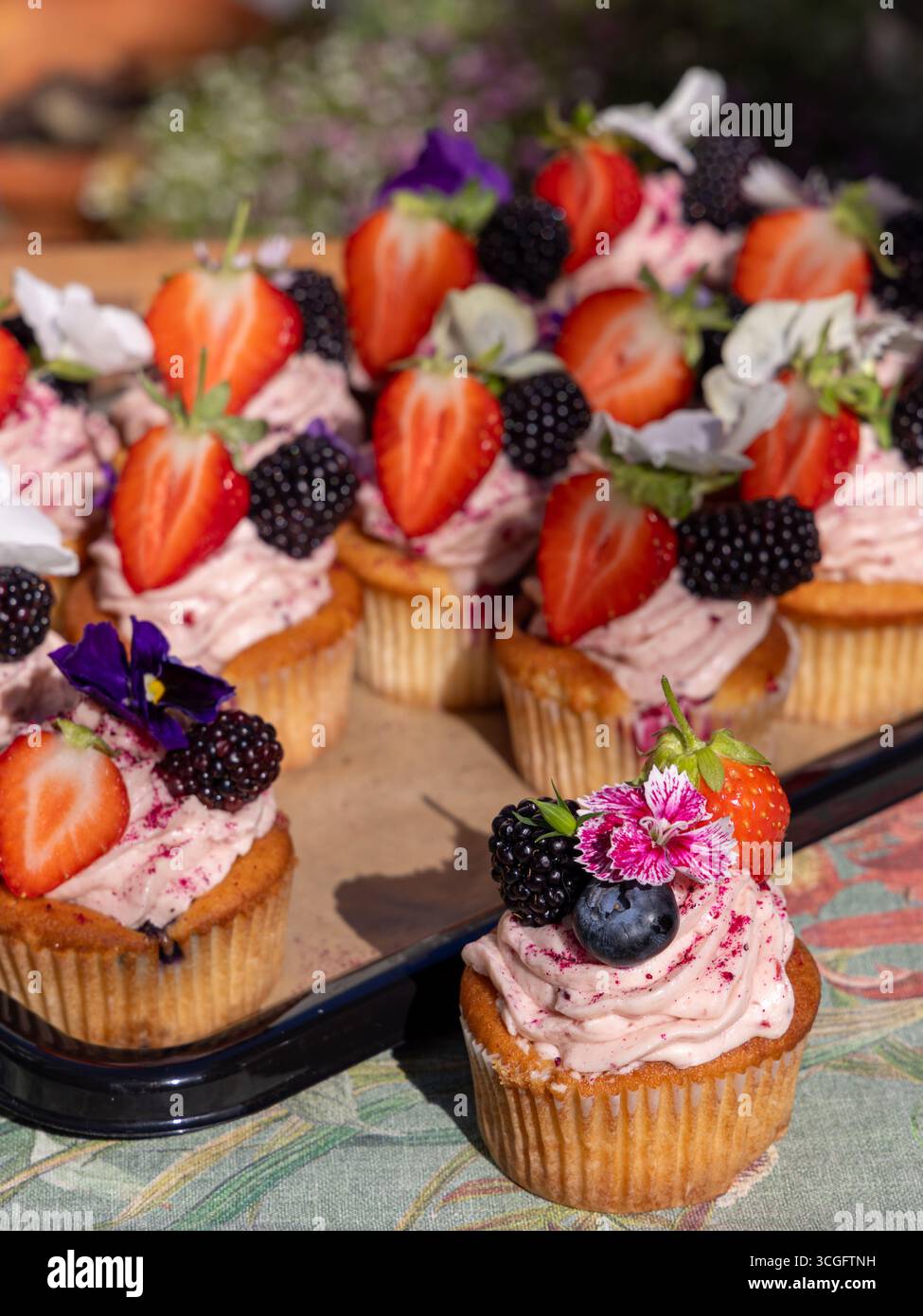 Peggy Porschen Sommer-Beeren-Cupcakes im Freien mit rosa Zuckerguss, frischem Obst und essbaren Blumen. Ein WhingEats-Gartenshooting. 🔍 47 K optimiert Stockfoto