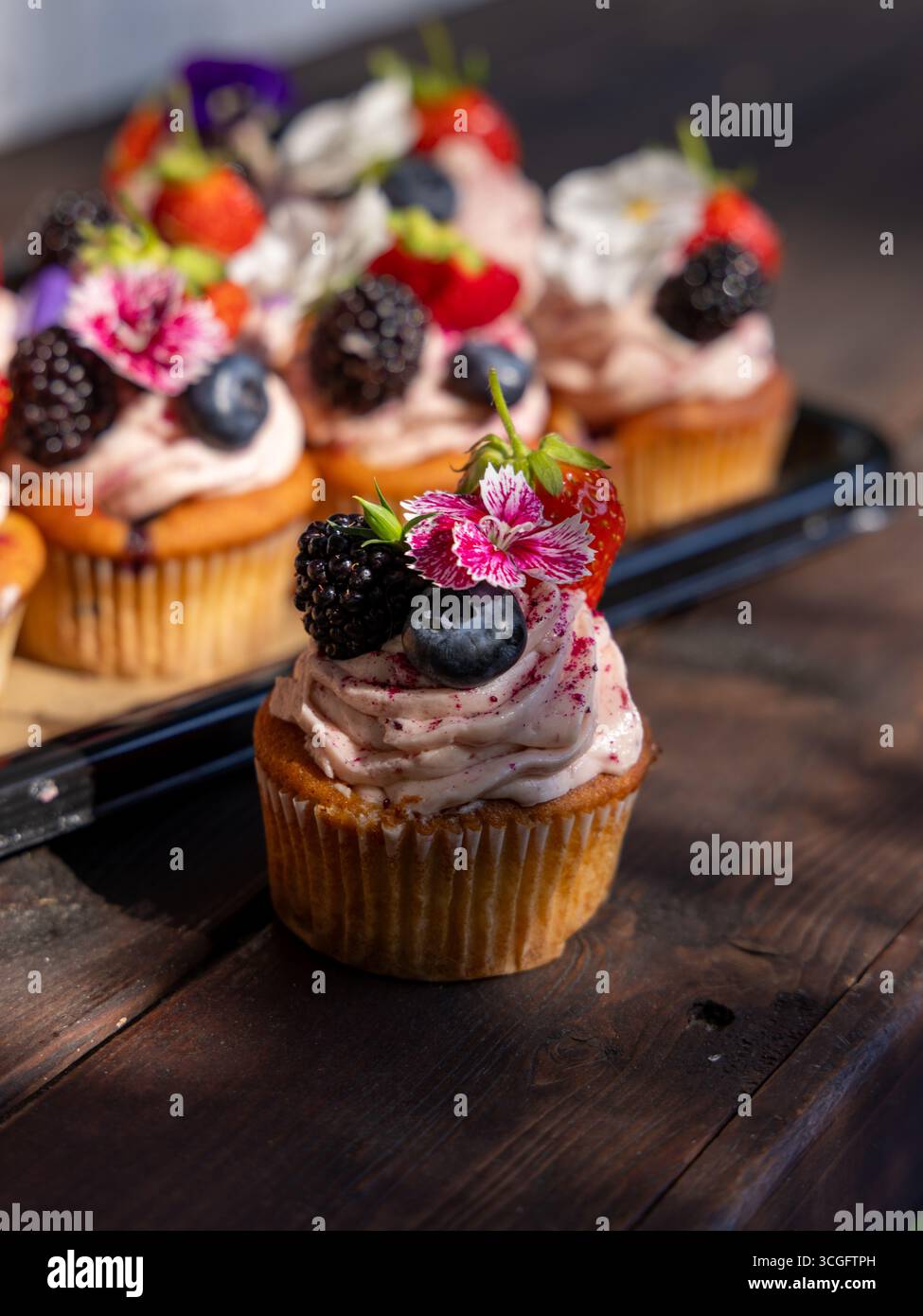 Sommerbeeren-Cupcakes von Peggy Porschen mit Beeren-Meringue-Zuckerguss, garniert mit Erdbeeren, Heidelbeeren, Brombeeren und essbaren Blumen. 🔍 47 Stockfoto