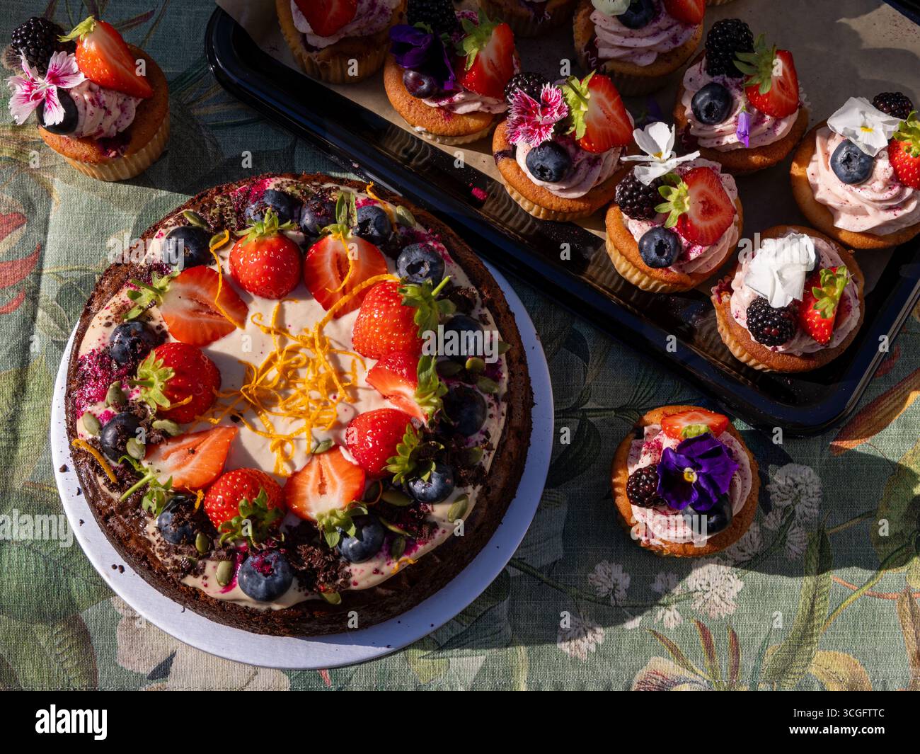 Tim Spectors Zoe-freundlicher Karottenkuchen mit Peggy Porschen Sommerbeeren-Cupcakes, dekoriert mit Früchten, Blumen und Nieseln auf floralem Tischtuch Stockfoto