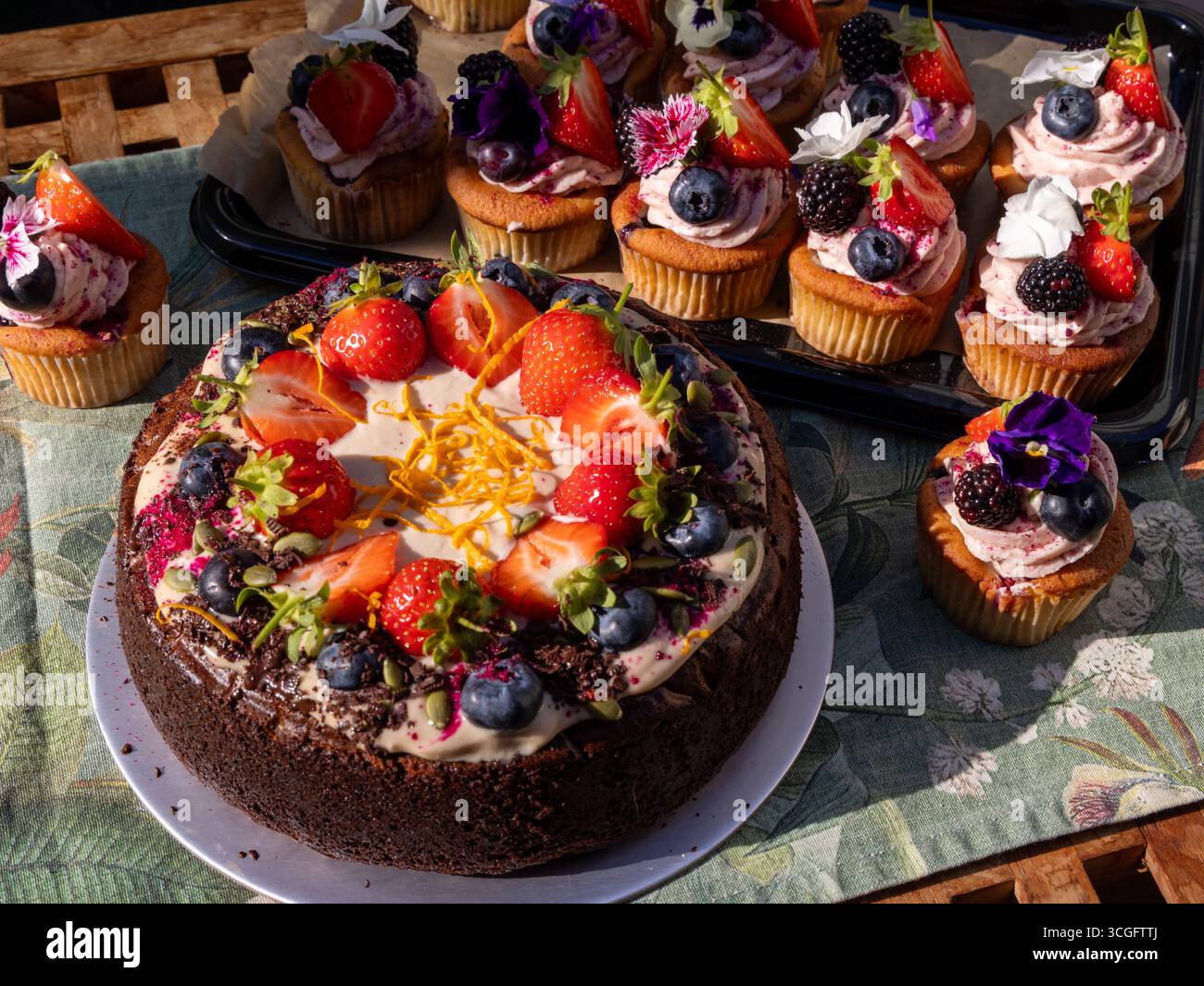 Tim Spectors Zoe-freundlicher Karottenkuchen mit Peggy Porschen Summer Berry Cupcakes, mit Früchten und Blumen auf gemusterter Tischdecke dekoriert. Stockfoto
