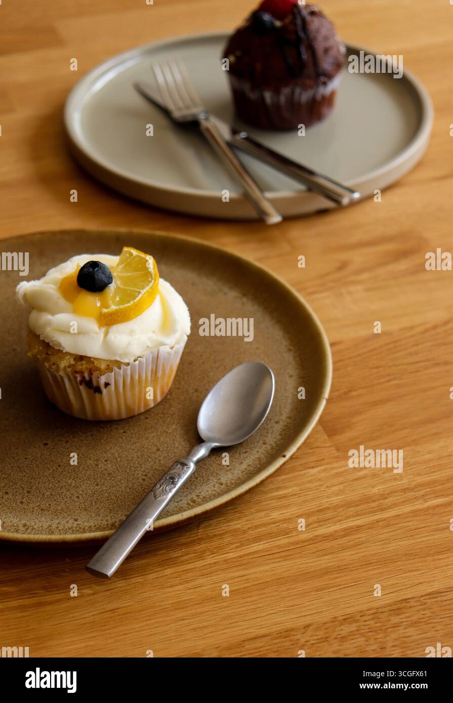 Ein köstlicher Vanille-Cupcake mit weißem Zuckerguss, einer Heidelbeere und einer Zitronenscheibe auf einer rustikalen Keramikplatte mit einem Löffel, mit einem zweiten Cupcake Stockfoto
