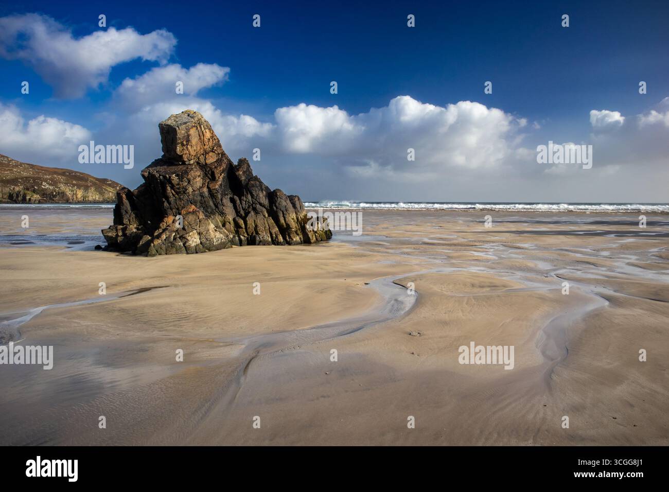 Die Felsformation steht an einem Sandstrand, umgeben von Gezeitenwassermustern unter einem tiefblauen Himmel mit verstreuten Wolken und Wellen in der Ferne Stockfoto