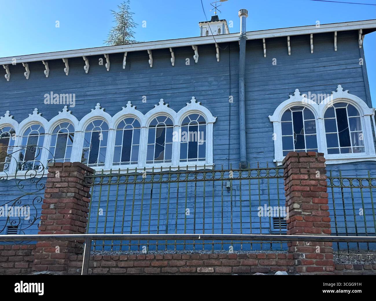 Äußere eines blauen Holzgebäudes mit dekorativen, weiß getrimmten Bogenfenstern, Blick von der Straße aus hinter einem Backstein- und Eisenzaun. - Smartphone-aufgenommenes Stockfoto