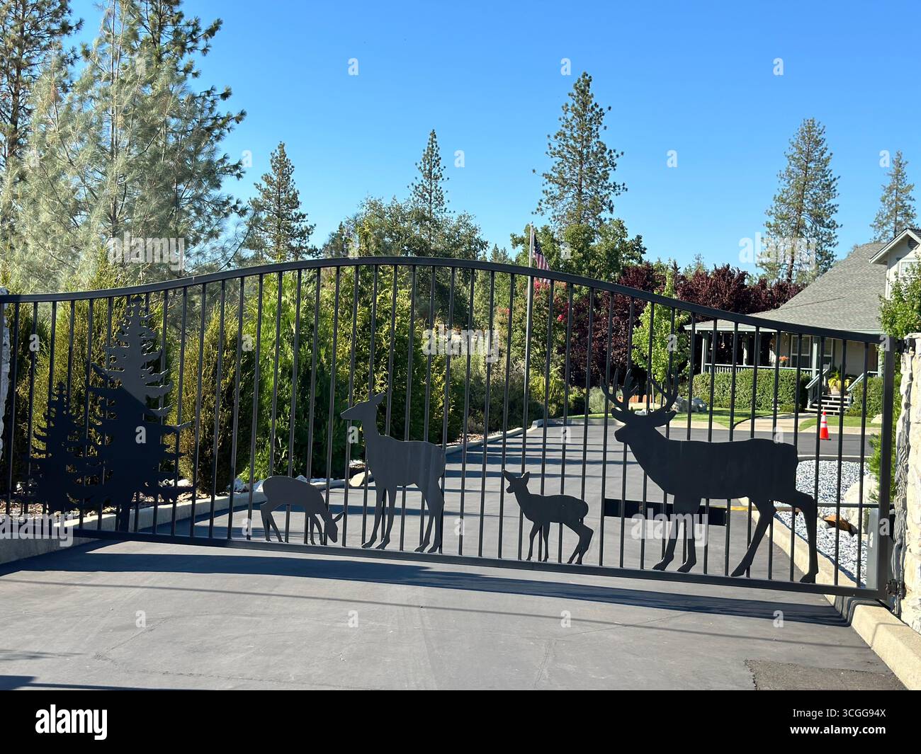 Ein privates Wohntor mit dekorativem Metalldesign aus Hirschen und Bäumen, mit einem Haus und einem begrünten Hof im Hintergrund. - Smartphone-aufgenommenes Stockfoto