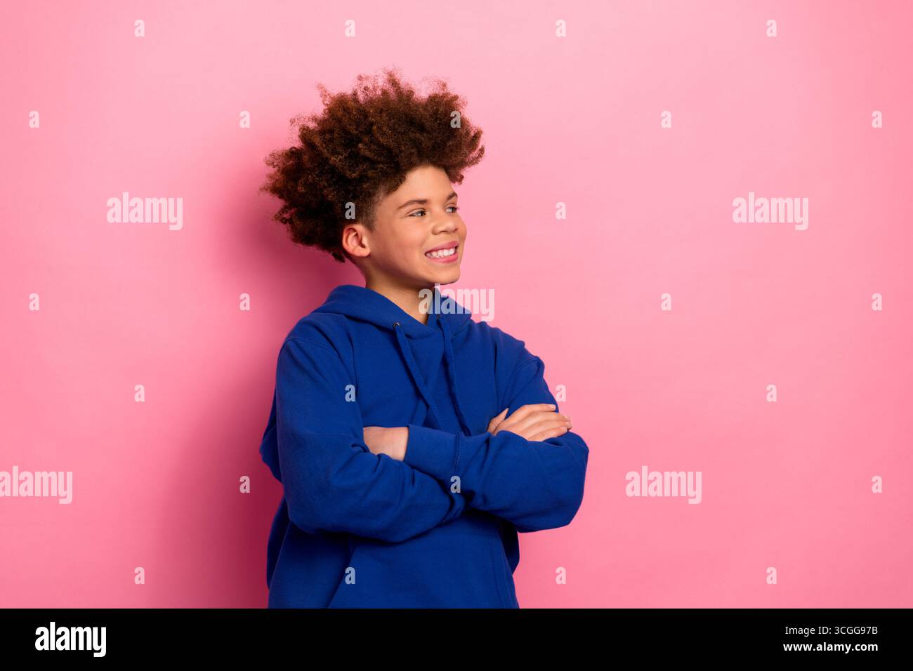 Fröhlicher Junge im lässigen blauen Hoodie mit überkreuzten Armen auf rosa Hintergrund, der Glück und stilvolle Jugendlichkeit darstellt Stockfoto