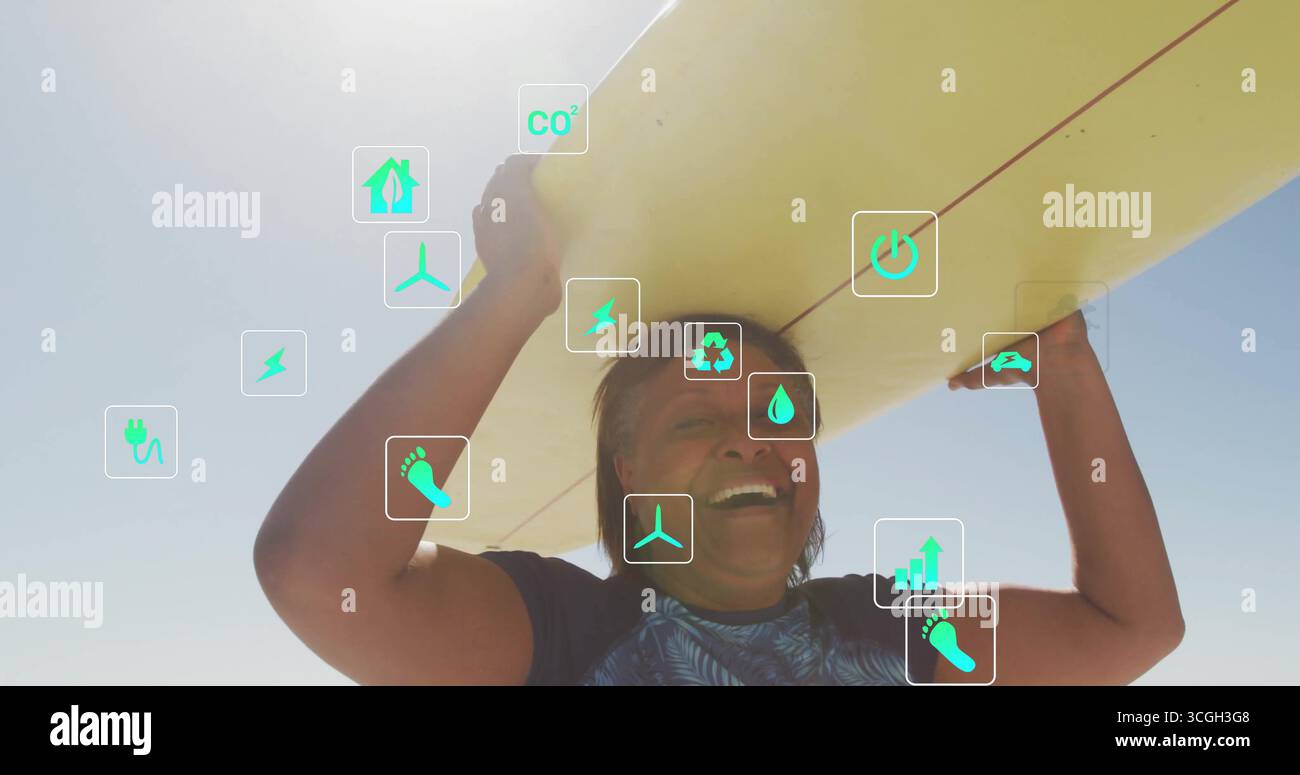 Lächelnde Frau im Badeshirt, die am Strand ein gelbes Surfbrett über dem Kopf hält, mit grünen Öko-Ikonen Stockfoto