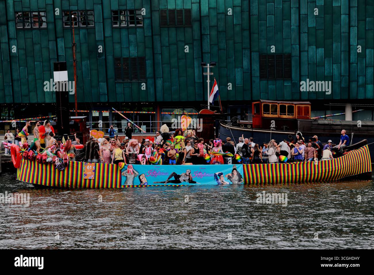 Das lange Regenbogenparadeboot mit Darstellern fährt während der Pride Amsterdam Canal Parade in Amsterdam, Niederlande, an einer grünen Kupferfassade vorbei. Stockfoto