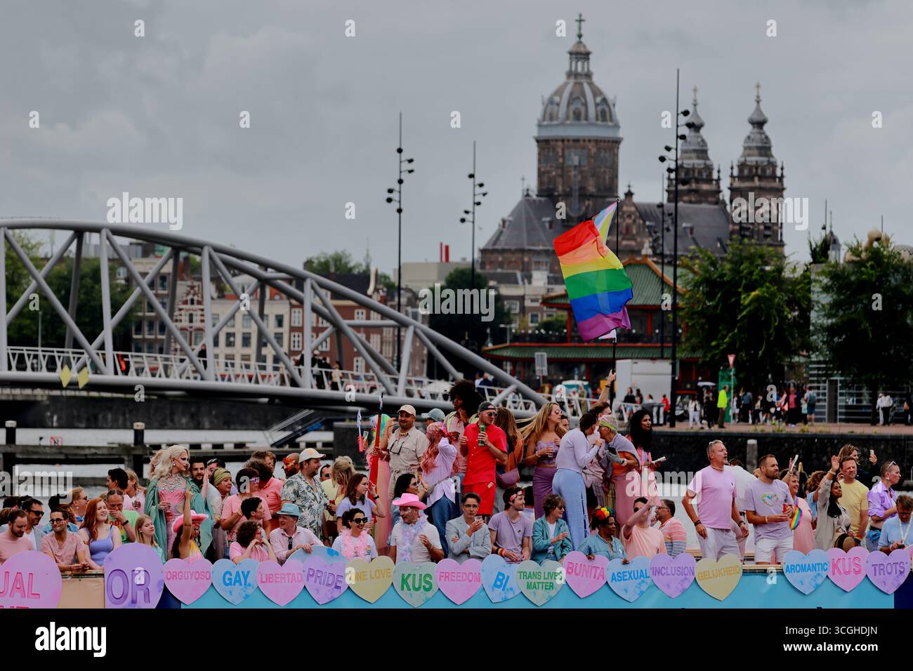 Ein Paradeboot mit Anhängern in pastellfarbenen Outfits und einer Regenbogenfahne passiert während des Pride Amsterdam in Amsterdam, Niederlande, eine Brücke mit der Skyline der Stadt. Stockfoto