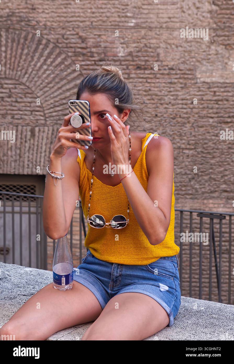Ein gebräuntes, attraktives Mädchen in gelber Weste und Jeansshorts sitzt auf einer niedrigen Wand und benutzt ihr Handy, um ihr Make-up zu überprüfen Stockfoto