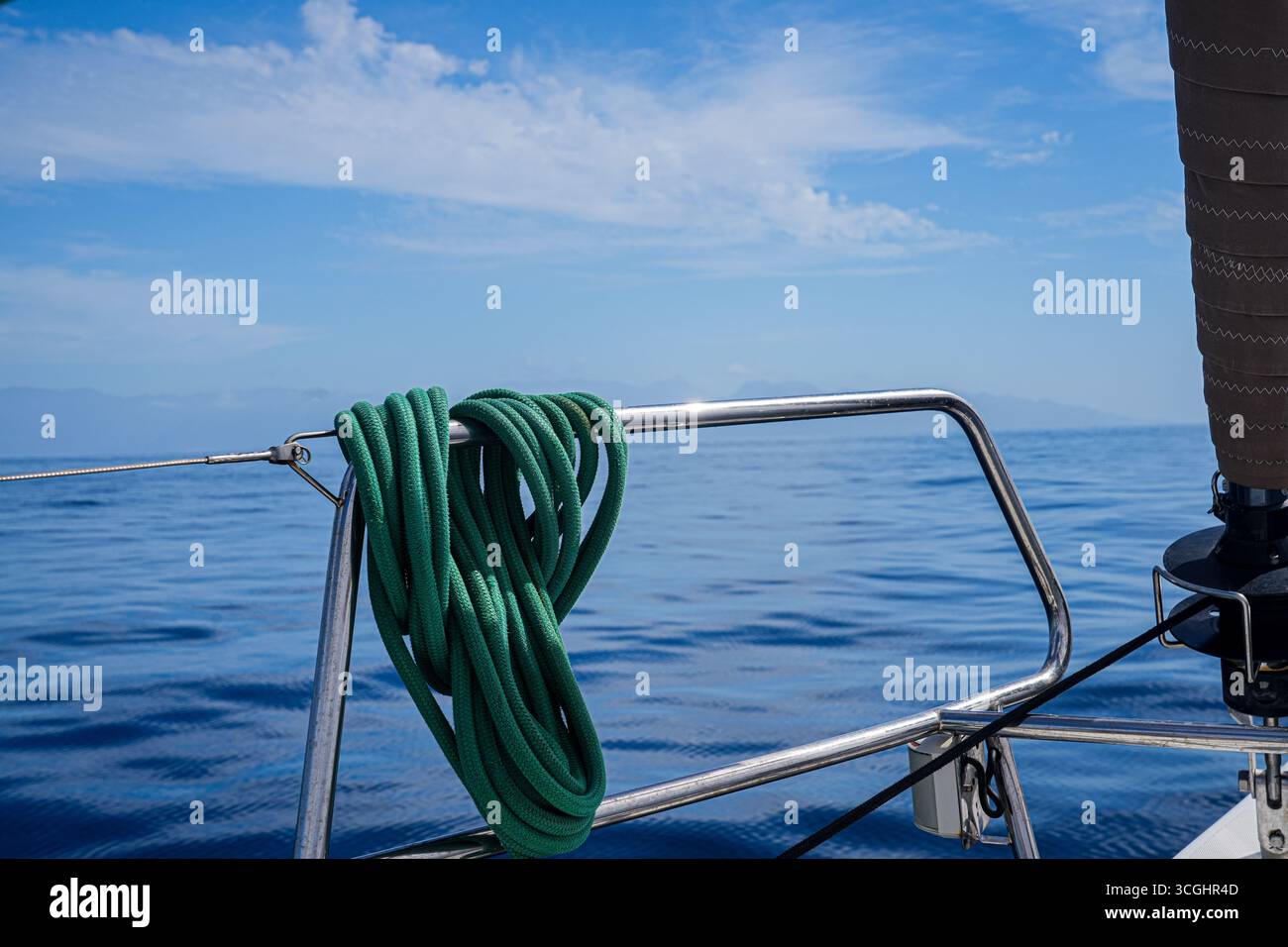 Gewickeltes grünes Seil auf einer Segelbootschiene über einem ruhigen blauen Meer; rostfreie Beschläge und Rollausrüstung sichtbar, nautische Reise, keine Leute. Stockfoto