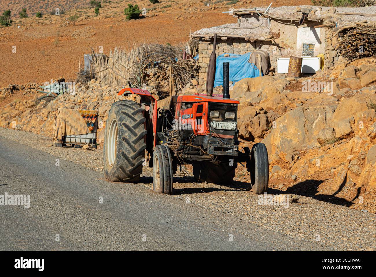 Alter roter Traktor, der an einer ländlichen Straße mit Steinhäusern und felsigem Gelände in der marokkanischen Landschaft geparkt ist Stockfoto