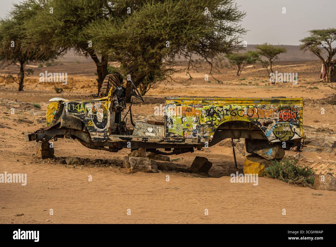 Verlassener, bemalter Lkw-Rahmen in der marokkanischen Wüste mit Graffitis, Akazienbäumen und trockener Landschaft im Hintergrund Stockfoto