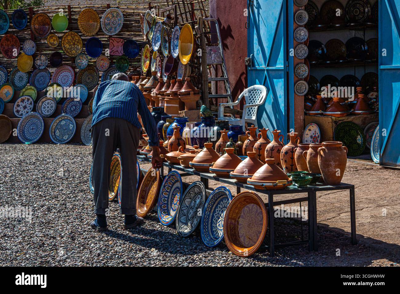 Ein einheimischer Mann arrangiert traditionelle marokkanische Tagine-Töpfe und bunte Keramikplatten auf einem Töpfermarkt im Freien in Marokko. Stockfoto