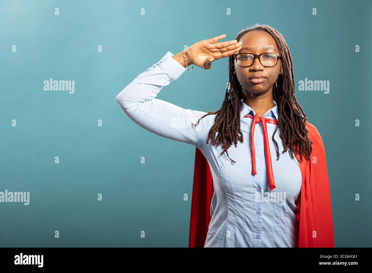 Ernste schwarze Heldin steht und respektvollen militärischen Gruß gibt. Die junge afroamerikanische Frau in rotem cape und blauem Hemd schlägt kraftvolle Posen und strahlt Vertrauen im Studio aus. Stockfoto