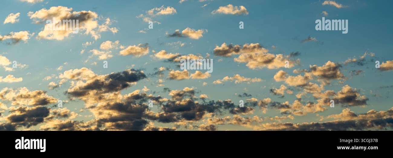 Kleine verstreute Wolken ziehen über einen klaren blauen Himmel, beleuchtet von den goldenen Tönen der untergehenden Sonne. Stockfoto