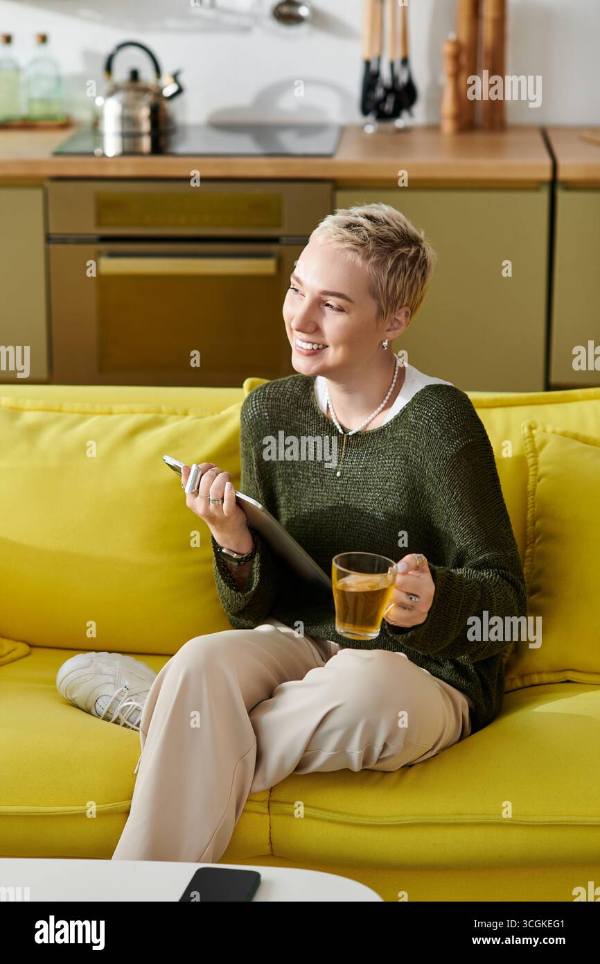 Eine schöne junge Frau mit kurzen Haaren lächelt, während sie bequem auf einer leuchtend gelben Couch sitzt. Stockfoto
